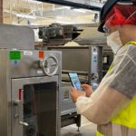 Wartungsperson mit Schutzkleidung prüft über ein Smartphone mit Inspect AR-App eine Maschine in der Lebensmittelproduktion in der Nestlé-Fabrik Wangen